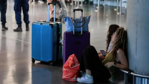 Two women sit on the floor of an airport, leaning against a concrete pole, their suitcases nearby
