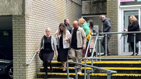 Mabli's family left court holding a soft toy. Two women wearing black dresses and light coloured cardigans walk down the stairs. One is holding a soft toy in her right hand which is also being held by a man who is bald and wearing beige trousers and jacket and a black top. Six other family members are visible walking behind them wearing coats. 