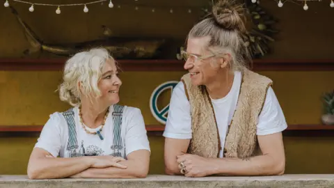 Camp Bestival Rob da Bank on the right leaning on a shelf looking at Josie da Bank on the left who is doing the same pose and looking back at him. The pair are smiling.