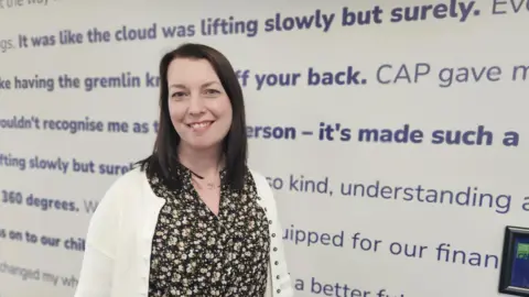 Aisha Iqbal/BBC A woman with dark shoulder-length hair, wearing a floral dress and a white cardigan. She stands next to a white wall covered in blue writing, all positive slogans and quotes like "the cloud was lifting, slowly but surely".
