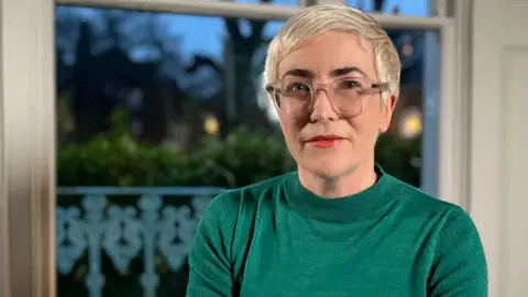 Alice Sullivan sits in a room looking at the camera. She has short platinum blonde hair, glasses and bright lipstick. She's wearing an emerald green jumper.