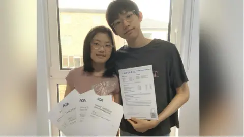 A family photo Ariel posing for a picture with her brother Elias. They are holding her GCSE results and his unconditional offer from Imperial College London. Both of them are wearing glasses.