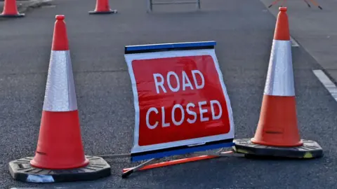 Two red and white traffic cones sit either side of a red and white sign that reads 'Road Closed'