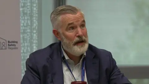 A man with white beard and hair sits at a table. He is wearing a dark blue suit and a lightly coloured checked shirt. 