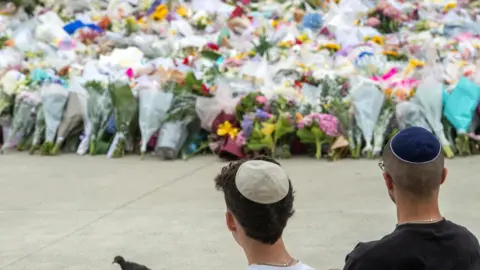 REUTERS/Jeremy Piper Members of the Jewish community sit near the floral tribute at Bondi Beach to honour the victims of a mass shooting targeting a Hanukkah celebration on Sunday at Bondi Beach, in Sydney