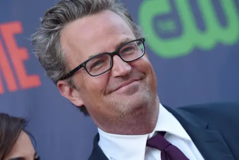 Actor Matthew Perry smiles in a dark suit and purple tie while arriving to a party 