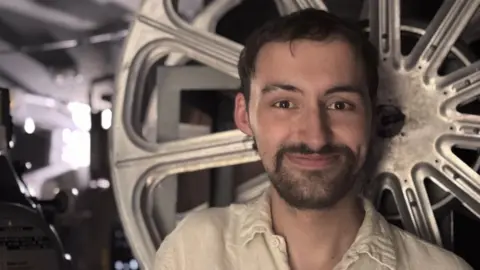 Patrick smiles at the camera, in front of a large wheel that holds film in the projection room. He has dark hair and a short dark beard, and wears a white loose shirt. The room behind him is dark and full of machinery