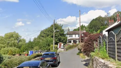 A narrow road with two cars on the left hand side in the foreground and a white house with a brown roof in the background