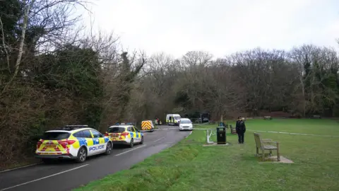 Police cars are seen parked along the side of the road next to a green space where there are park benches and trees that have no leaves on them. A lone police officer is seen facing the camera in front of some police tape.