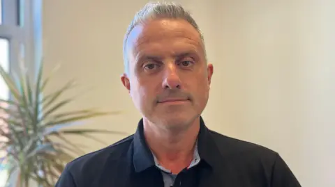 A man wearing a black collared shirt with a plant and plain wall behind.