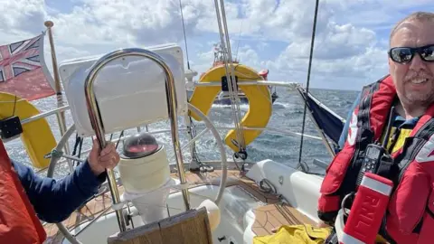 Two people wearing life jackets on board a boat with a Union Jack at the rear and an RNLI lifeboat behind it on the sea