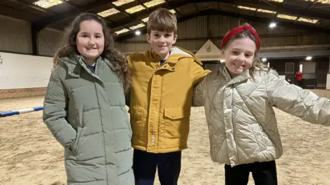 Leanne Rinne/BBC Evalyn has brown hair and is wearing a grey coat, Leo has brown hair and is wearing a yellow coat, and Eadie is wearing a red headband and a gold puffer coat. The trio are pictured at the East Park Riding for the Disabled Group in Lingfield, Surrey.