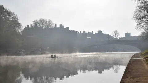 Isaac Savage The River Ouse taken in the morning, with mist over the water and it is just getting light. Two people are rowing in the middle of the water and just their silhouettes are visible.