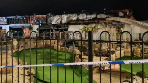 A closer view of the damage to the sign outside the amusement arcade. The images on the sign have burnt away leaving black scorch marks and silver metal behind. A mini-golf course decorated with dinosaurs is in front of the building.