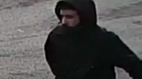 West Yorkshire Police A man whose face is partially obscured by a black hooded coat. The image is grainy.