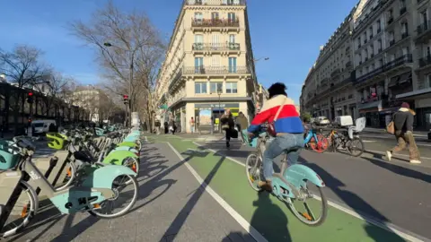 A cyclist in a red, white and blue coat is driven by a green bicycle tape in Paris next to a series of bicycles to rent