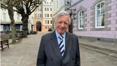 Deputy Sir Philip Bailhache is pictured outside the States Assembly. He is wearing a grey blazer and blue shirt. 