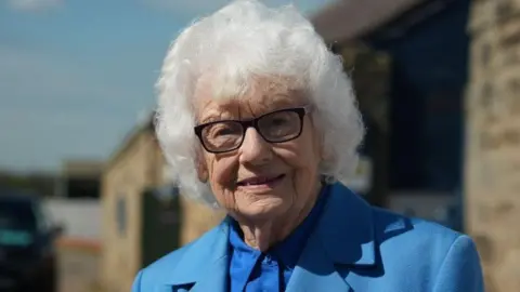 Vera Shearer smiles at the camera. She has thick curly white hair, black glasses and is wearing a blue shirt and jacket.