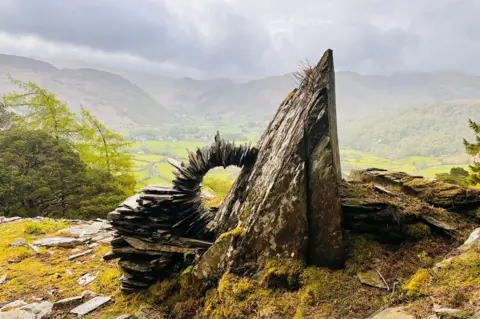 Clive Rowlandson Flat slate and stones piled on top of each other in a curve against a rock face