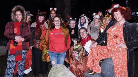 A group of women, some dressed as foxes and some just wearing orange, smiling at the camera. It's dark and there's a tree behind them.