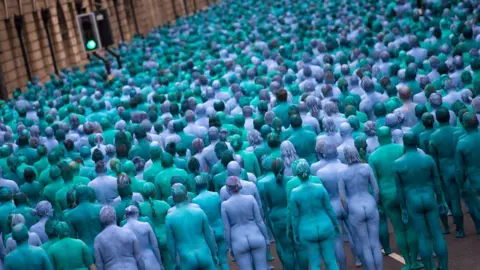The backs of blue, naked participants all standing facing away from the camera. A traffic light on green pokes out above the sea of bodies. Part of the stone building of the Guildhall can be seen in the top left hand corner.