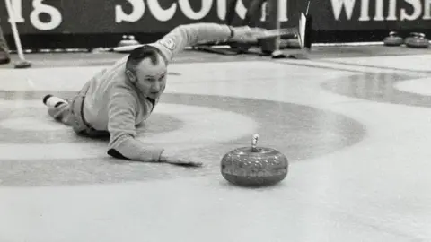 Hammy McMillan lying on his stomach on the ice with his right arm resting on the ice, having just thrown the curling stone- his left arm is held in the air
