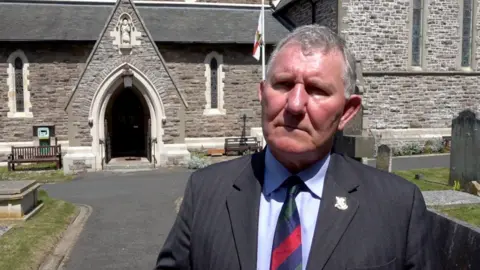 A man in a suit stood in a church graveyard. There is a flag in the background.