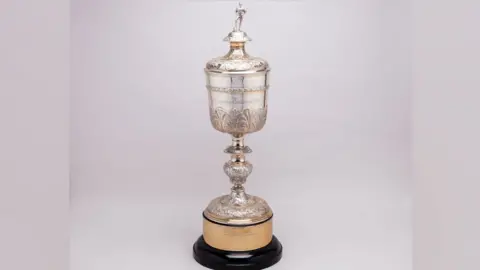 BUDDS The Littlewoods League Cup trophy, a solid silver chalice-like cup. It has a lid on which stands the figure of a male footballer with his hands on his hips and his left leg resting on a ball. The cup is ornate with a leaf pattern around its stem and base. It is resting on a gilded base, which in turn is resting on a wooden base. 