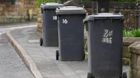 Three black wheelie bins have been left out for collection in front of low stone wall on a kerbside. 