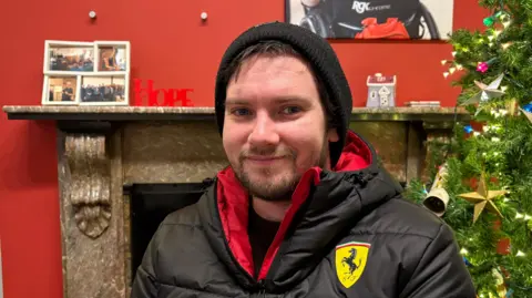 BEN SCHOFIELD/BBC Andrew Rayner looking directly down the camera and smiling. He is inside, seated in front of a fire surround and a Christmas tree, on the right of the image. He is wearing a black woollen hat, from which a small amount of fringe hair is poking, and has dark stubble on his chin. He is wearing a thick, black coat, whose lining is red. The coat also has a yellow insignia with a prancing black pony on. The walls in the room are a deep
