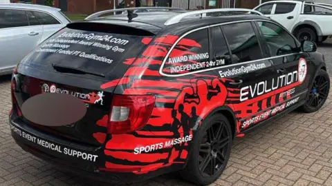 Micky Donkin The four-door car is black with red decal displaying information about the Evolution Fitness gym, including an email address and website url.