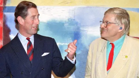 The then-Prince of Wales standing next to and speaking to David Hockney in front of one of his brightly-coloured paintings in 1994