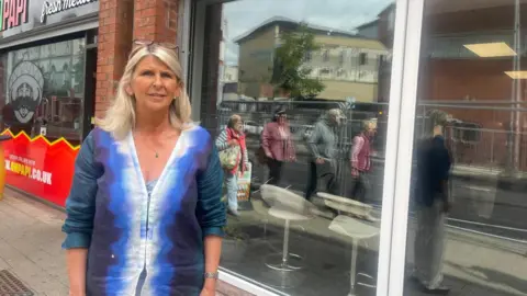 a lady in a blue jacket stands in front of a row of shop windows. she has long blond hair and has sunglasses atop her head