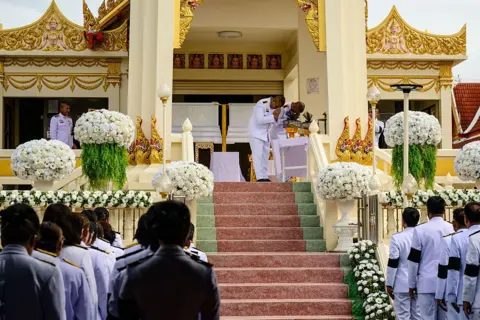 AFP Mourners gather by a crematorium decorated with white floral arrangements as uniformed attendees stand on either side of a staircase. At the top of the steps, two officials stand beside white coffins.