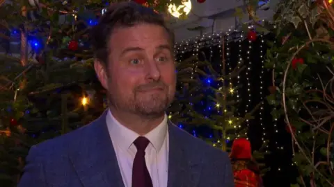 A man in a blue suit and red tie is surrounded by Christmas trees and fairy lights.