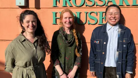 Three women stand smiling at camera with backs against the wall of a public building