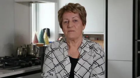 Katie is sat in a kitchen. She is wearing a black and white patterned blouse and is looking into the camera.