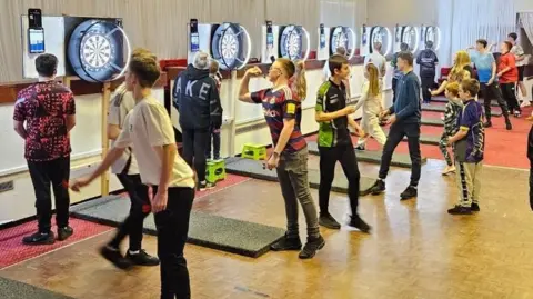 Simon Tate About 20 children of all ages are playing darts. They are in groups of three or four in front of seven darts boards 