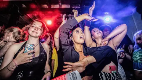 Getty Images Three young women dancing in a Manchester nightclub with a crowd and flashing red and blue lights behind them