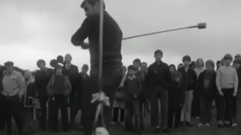 Still from BBC Nationwide 1972. A man about to hit a ball (knurr) with a stick (spell), watched by a crowd.