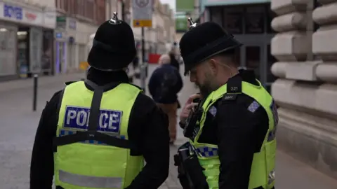 Two police officers in yellow high vis uniform vests are standing near each other on Hull's High Street. PC Gareth Bielby on the right is talking into his police radio. Sergeant Lee Waller, on the left, is looking straight ahead.