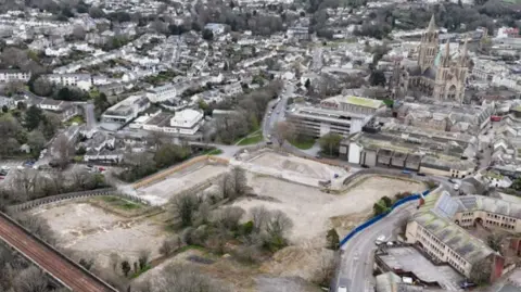 An aerial view shows a large cleared construction site in Truro city centre, surrounded by roads and buildings, with a railway line running along one edge. A cathedral with tall spires dominates the scene nearby, with housing estates and green countryside stretching into the distance.