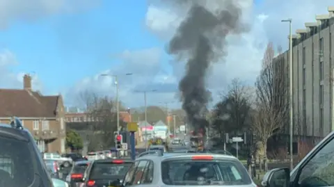 A row of cars stopped at a red traffic light. In the background a lorry is on fire. A large plume of black smoke is billowing from the vehicle and rising up to a blue sky