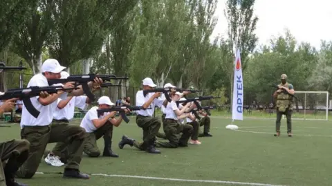 Yunarmia branch of the Zaporizhzhya region A line of around ten boys stand in a football pitch, kneeling and aiming a gun. They are wearing khakis and white t-shirt. An adult dressed in army camos and a bullet proof vest looks on. he is wearing a balaclava and is armed with a large gun.