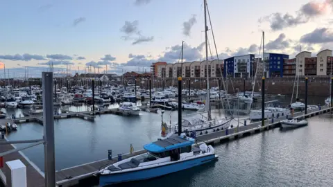 St Helier marina filled with moored boats and yachts at dusk. Blocks of flats visible in the distance.