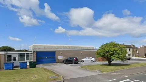 A Google Street View screenshot of the entrance to Distington Ambulance Station: a low, concrete building, with a large garage-style entrance painted in blue. In the foreground, to the right, is a tree standing on a lawn. To the left, another area of green, with industrial bins in front of a white door and window.