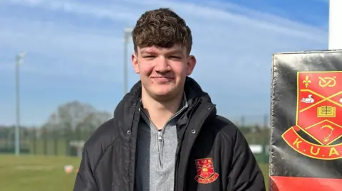 Spencer has brown hair and is wearing a grey top and black coat. He is holding a white and red rugby ball. He is standing on a green rugby pitch with a rugby post behind him, that has a padded material over it with the Keele University logo on a red background