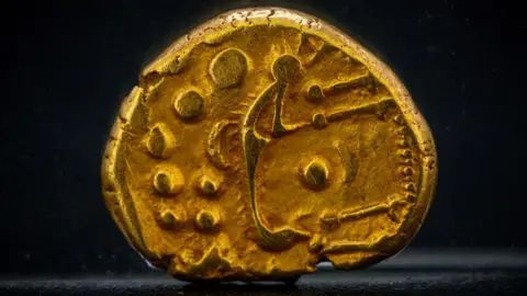 Fountain Media A close-up of a gold Iron Age coin. It is a rounded oval in shape and has dots and shapes struck on it.