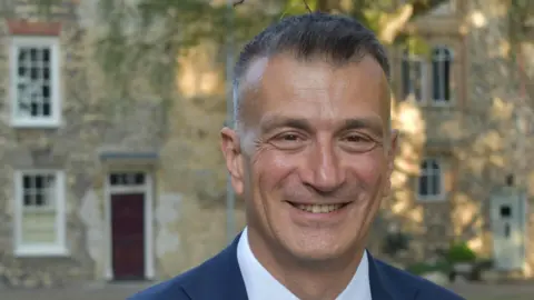 Eastern Education Group Dr Nikos Savvas smiles at the camera. He has short dark hair with some grey in it. He wears a navy suit with a white shirt. He is standing outside and some homes can be seen behind him.