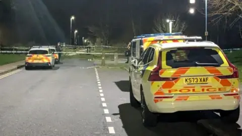 Three police cars and a police van parked on either side of an empty road. Police officers and a cordon can be seen in the distance